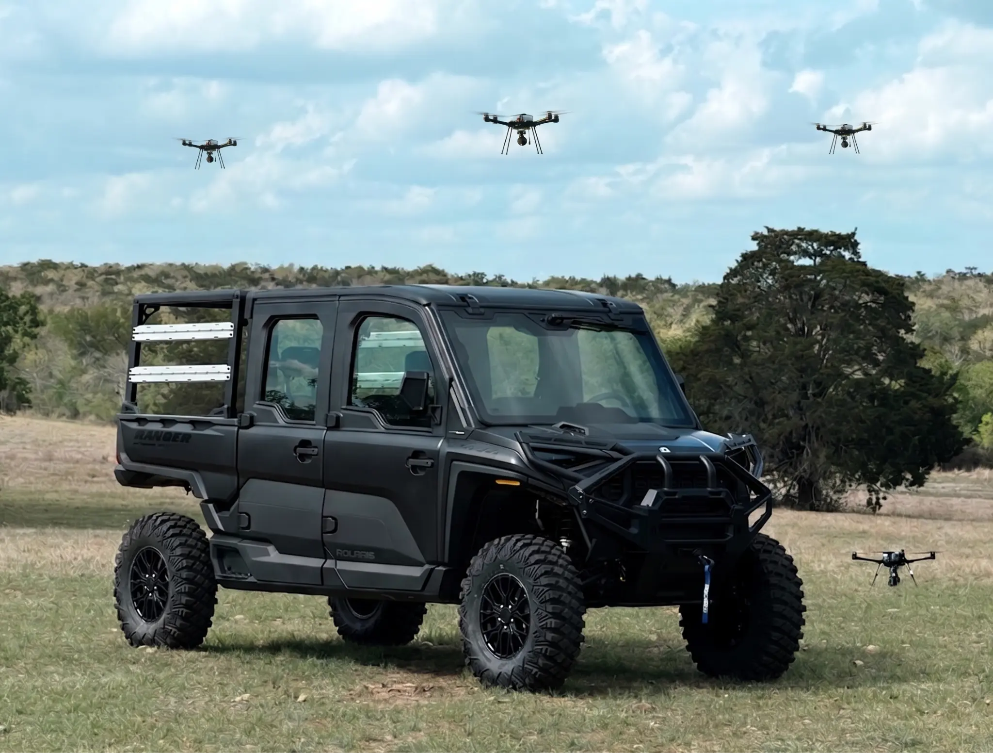Off-road ground vehicle with multiple drones flying above it in a field.
