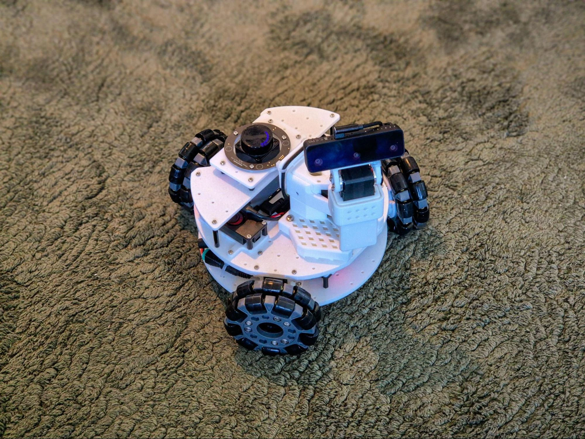 The LeKiwi mobile robot on a green carpet, with its pan-tilt depth camera, 360° LiDAR, and omni wheels
