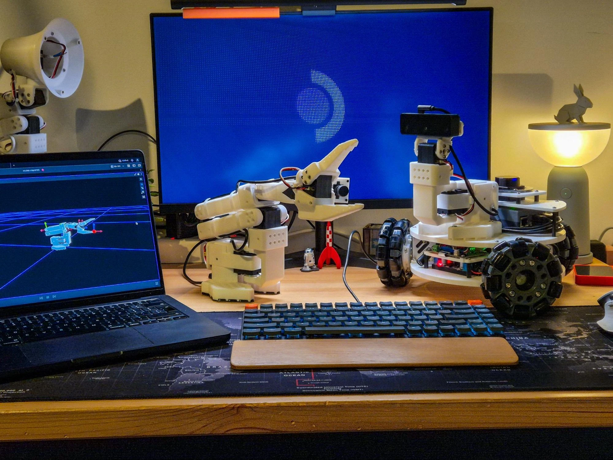 Desk setup with an SO-101 robot arm, a LeKiwi mobile robot, and Foxglove showing the arm on a laptop
