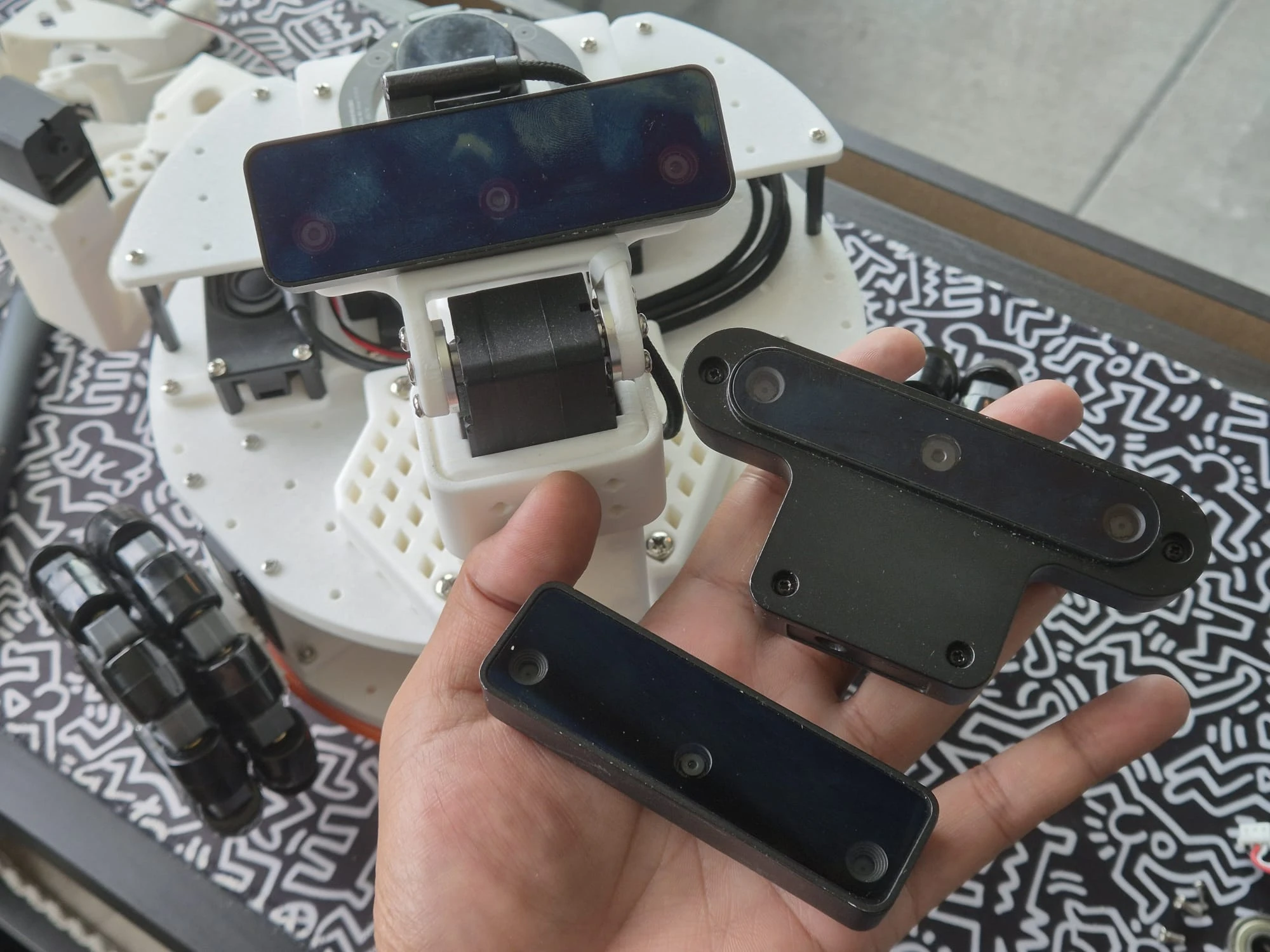 Hand holding two Luxonis OAK cameras, with a third already mounted on the robot's pan-tilt mechanism in the background